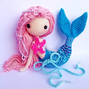 Può includere: Una bambola sirena all'uncinetto con capelli rosa, coda blu e una stella marina rosa. La bambola indossa una collana di perle e ha un nastro blu chiaro legato alla coda.