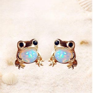 Puede incluir: Un par de pendientes dorados con forma de rana. Cada pendiente presenta una piedra de color azul claro, similar al ópalo, para el vientre de la rana y ojos negros. Los pendientes están sobre un fondo arenoso con una concha.