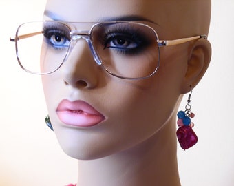 May include: A silver-framed pair of clear aviator glasses with a gold nose bridge. The glasses are worn by a mannequin with pink lips and dark eye makeup. The mannequin is also wearing dangle earrings with pink, blue, and green beads.