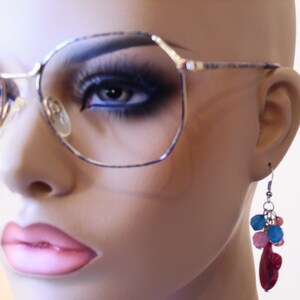Peut inclure: Une paire de lunettes argentées avec une monture géométrique et des verres teintés bleus. Les lunettes sont portées par un mannequin avec des lèvres roses et une paire de boucles d'oreilles pendantes avec des perles rouges, roses et bleues.