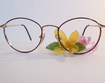 May include: Round eyeglasses with a gold-colored frame and tortoise shell accents on the rims. The glasses have clear lenses and black temples. A small arrangement of yellow and pink flowers rests beneath the glasses.