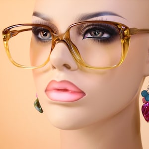 May include: A mannequin head wearing large, square, amber-colored eyeglasses. The glasses have a thick frame and a classic, retro style. The mannequin also has long eyelashes, pink lipstick, and dangling earrings with colorful beads and a leaf-shaped charm.