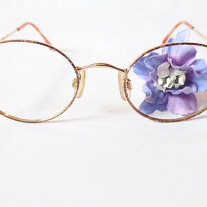 Può includere: Occhiali rotondi con montatura dorata, con un fiore viola visibile attraverso le lenti.