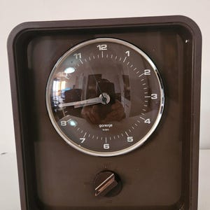 Può includere: Un orologio Gorenje kvarc di forma quadrata color marrone con quadrante circolare, numeri bianchi e lancette argentate. Sotto il quadrante si trova una manopola con segni per le impostazioni dell'ora. L'orologio ha una cornice in plastica marrone lucida.