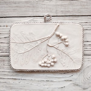 Puede incluir: Un relieve de yeso blanco de una rama de serbal con hojas y bayas. El relieve está enmarcado con un borde de cuerda y tiene un lazo en la parte superior para colgar.