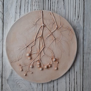May include: A round, peach-colored decorative plaque with a raised relief design of a plant with leaves and berries. The plaque is set against a weathered, white wooden background.
