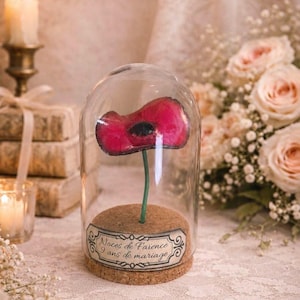 May include: A red poppy flower with a black center, encased in a glass cloche. The flower sits on a cork base with a label that reads "Noces de Faïence 9 ans de mariage". A candle and flowers are in the background.