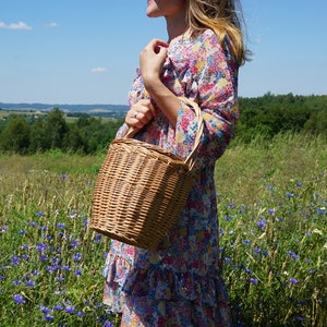 May include: A woman in a floral dress holds a woven wicker basket with a handle in a field of wildflowers.