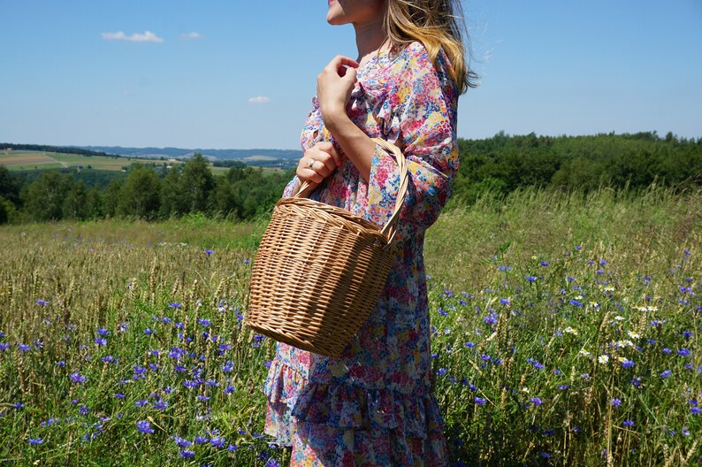 May include: A woman in a floral dress holds a woven wicker basket with a handle in a field of wildflowers.