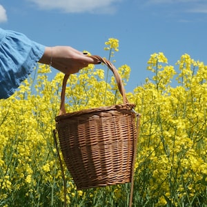 May include: A person holding a brown wicker basket with a handle in a field of yellow flowers.