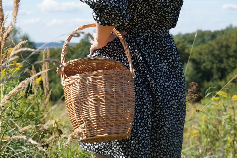May include: A woman in a black and white floral dress carries a woven wicker basket with a handle.
