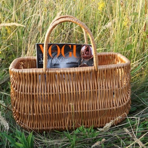 May include: A brown wicker basket with a handle, sitting in a field of tall grass. The basket contains a magazine with the word "Vogue" on the cover.