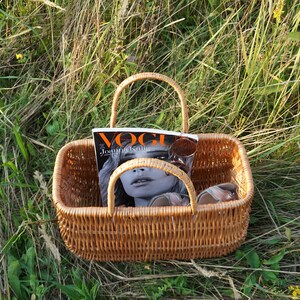 May include: A brown wicker basket with a handle, containing a magazine with the word "Vogue" on the cover, and sunglasses. The basket is sitting in a field of green grass.