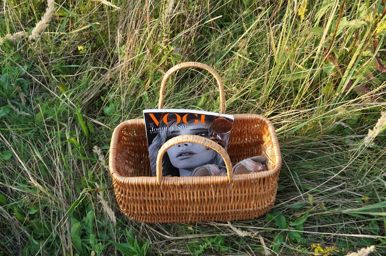 May include: A brown wicker basket with a handle, containing a magazine with the word "Vogue" on the cover, and sunglasses. The basket is sitting in a field of green grass.
