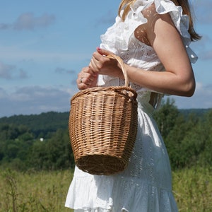 May include: A woman wearing a white dress holds a brown wicker basket with a handle. The basket is in focus, while the woman and the background are blurred.