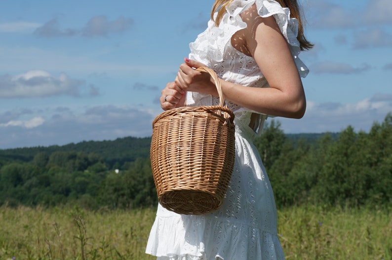 May include: A woman wearing a white dress holds a brown wicker basket with a handle. The basket is in focus, while the woman and the background are blurred.