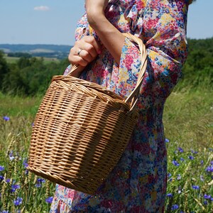 May include: A woven wicker basket with a handle, held by a person wearing a floral dress. The basket is brown and has a natural, rustic look.