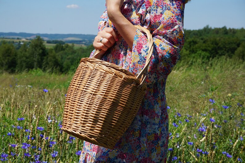 May include: A woven wicker basket with a handle, held by a person wearing a floral dress. The basket is brown and has a natural, rustic look.