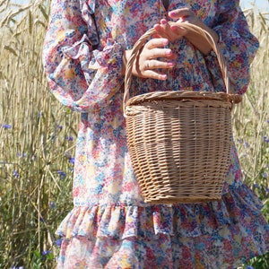 May include: A woman in a floral dress holds a wicker basket with a handle. The basket is woven with a light brown color. The woman is standing in a field of tall grass.
