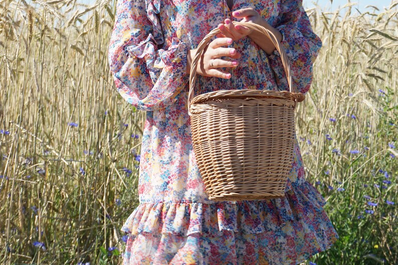 May include: A woman in a floral dress holds a wicker basket with a handle. The basket is woven with a light brown color. The woman is standing in a field of tall grass.