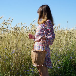 May include: A woman in a floral dress walks through a field of wheat, carrying a wicker basket.