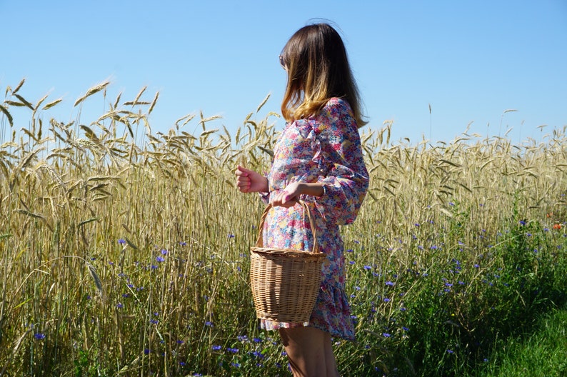 May include: A woman in a floral dress walks through a field of wheat, carrying a wicker basket.