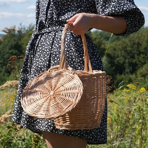 May include: A woman in a black and white floral dress holds a woven wicker basket with a round lid. The basket is made of natural materials and has a rustic look.