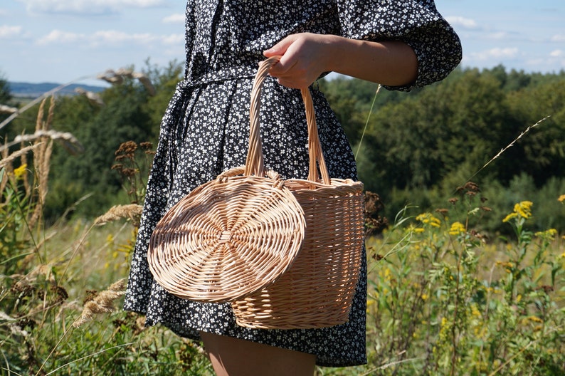 May include: A woman in a black and white floral dress holds a woven wicker basket with a round lid. The basket is made of natural materials and has a rustic look.