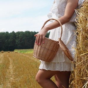 May include: A woman in a white dress holds a woven wicker basket in a field of golden hay bales.