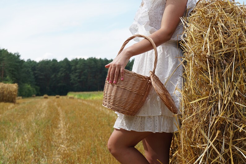 May include: A woman in a white dress holds a woven wicker basket in a field of golden hay bales.