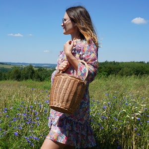 May include: A woman in a floral dress holds a brown wicker basket with a handle in a field of wildflowers.