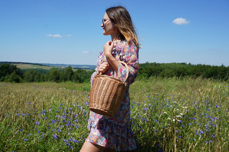 May include: A woman in a floral dress holds a brown wicker basket with a handle in a field of wildflowers.