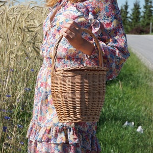 May include: A woman in a floral dress holds a brown wicker basket with a handle.
