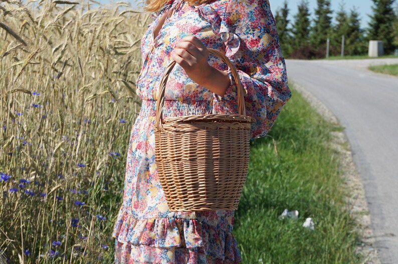 May include: A woman in a floral dress holds a brown wicker basket with a handle.