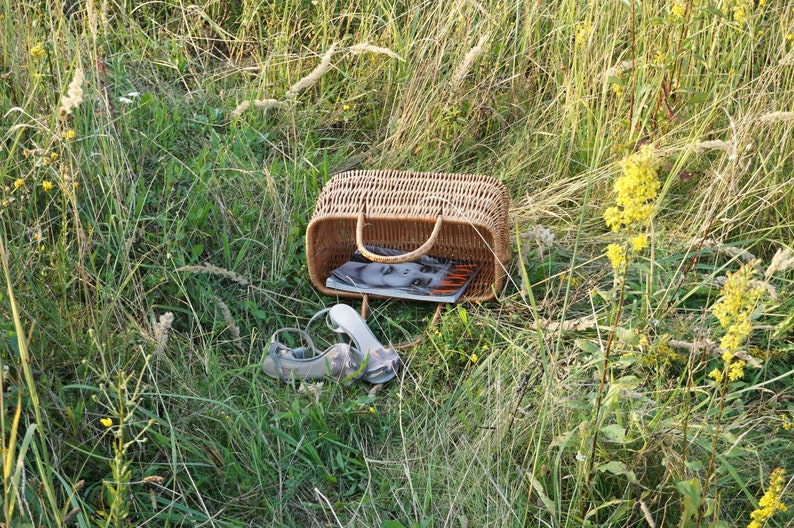 May include: A wicker basket with a magazine inside, sitting in a field of tall green grass with yellow wildflowers. Two gray sandals are lying on the ground next to the basket.