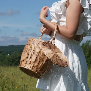 May include: A woman wearing a white dress holds a wicker basket with a lid. The basket is round and features a woven design. The woman is standing in a field with green grass and trees in the background.