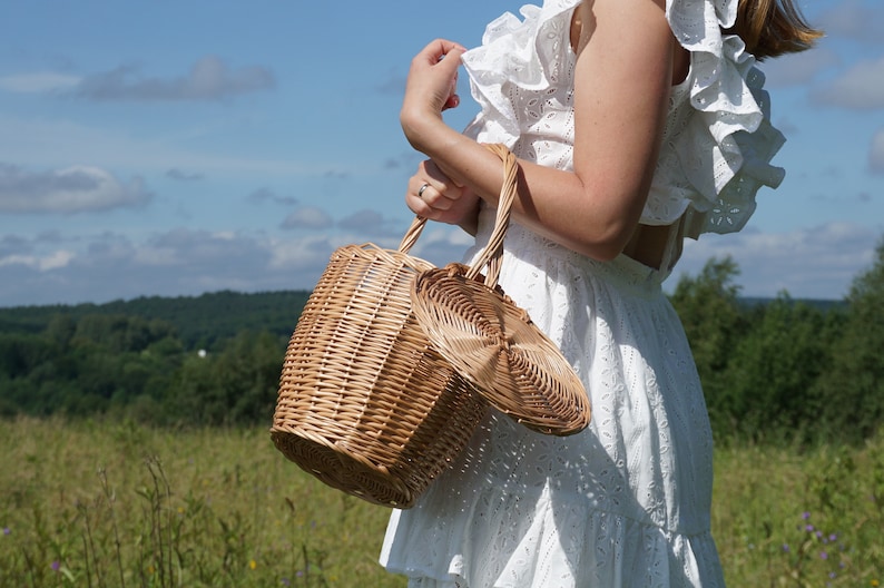 May include: A woman wearing a white dress holds a wicker basket with a lid. The basket is round and features a woven design. The woman is standing in a field with green grass and trees in the background.