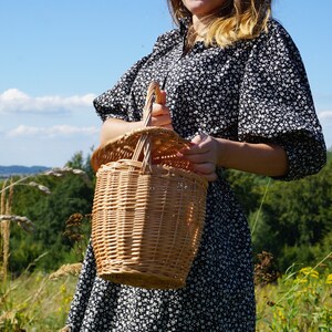 May include: A woman in a black and white floral dress holds a woven wicker basket with a handle.