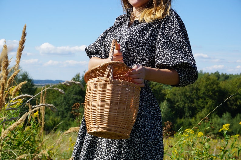 May include: A woman in a black and white floral dress holds a woven wicker basket with a handle.