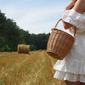 May include: A woman in a white, ruffled dress carries a woven wicker basket in a field of golden hay.