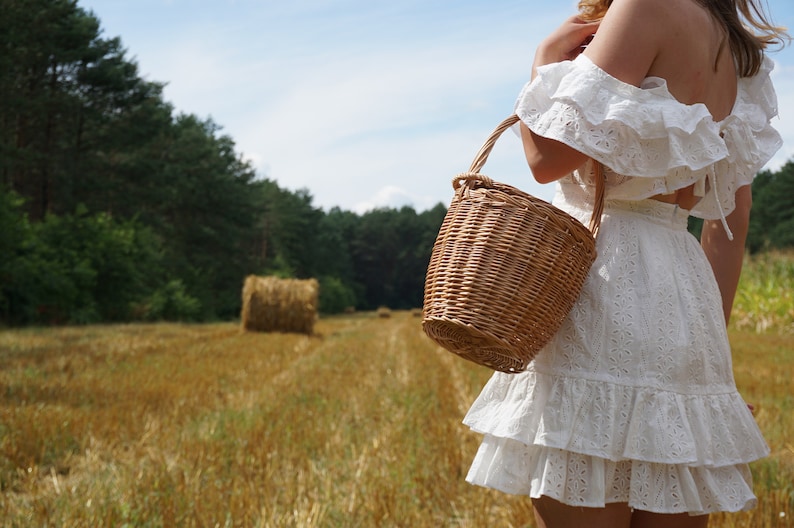 May include: A woman in a white, ruffled dress carries a woven wicker basket in a field of golden hay.