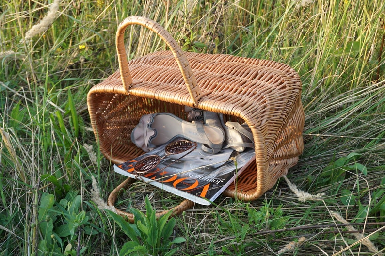 May include: A wicker basket with a brown handle, filled with a magazine, sunglasses, and sandals. The basket is sitting on a bed of green grass.