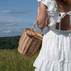 May include: A woman wearing a white eyelet dress with a ruffled neckline and a bow at the back is holding a woven wicker basket.