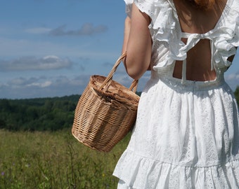 May include: A woman in a white eyelet dress with a ruffled neckline and a bow at the back is holding a woven wicker basket.