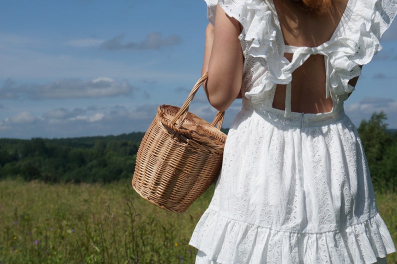 May include: A woman wearing a white eyelet dress with a ruffled neckline and a bow at the back is holding a woven wicker basket.