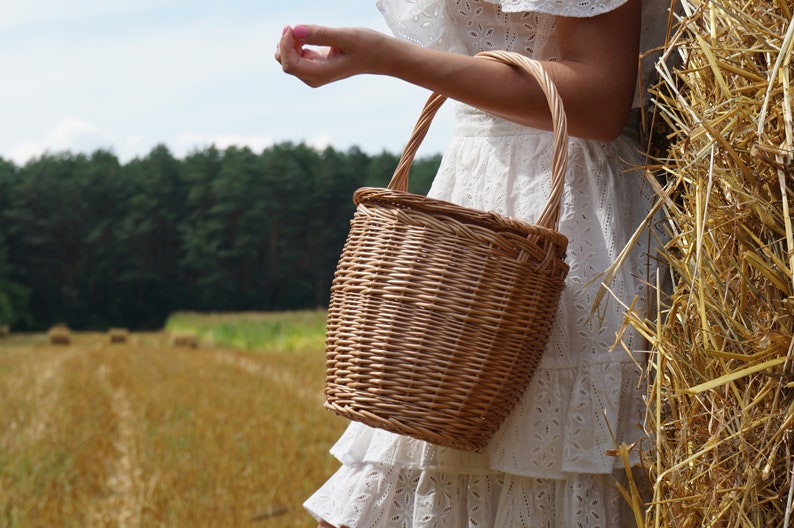 May include: A woven wicker basket with a handle, in a field of golden hay. The basket is light brown and has a natural, rustic look.