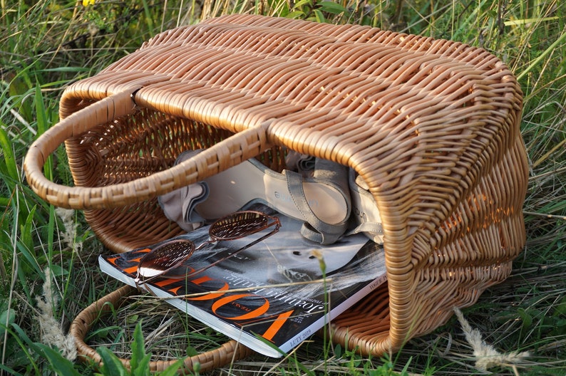 May include: A woven wicker basket with a brown stain. The basket is filled with a magazine, sunglasses, and a pair of sandals.