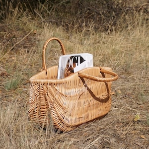 May include: A woven wicker basket with a magazine inside. The basket has two handles and is sitting on a bed of dry grass.