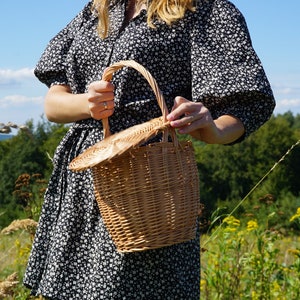 May include: A woman in a black and white floral dress holds a brown wicker basket with a lid. The basket has a woven handle and is in a field with green grass and yellow flowers.