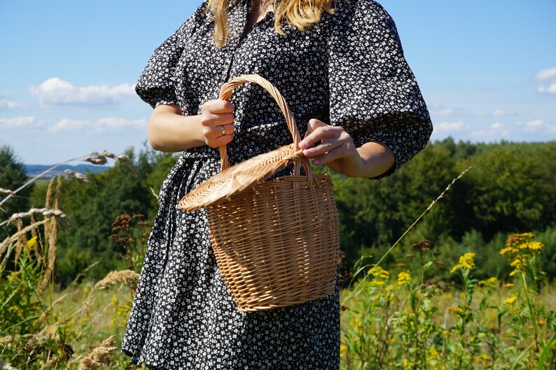 May include: A woman in a black and white floral dress holds a brown wicker basket with a lid. The basket has a woven handle and is in a field with green grass and yellow flowers.